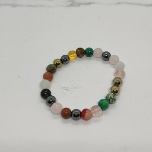 Könnte beinhalten: Ein Armband mit bunten Edelsteinperlen. Das Armband enthält Perlen in Rosa, Grün, Gold, Braun und Schwarz. Das Armband ist auf einem elastischen Band aufgefädelt.