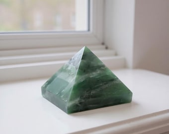 May include: A green pyramid-shaped decorative object. The pyramid is made of a translucent, green material with white veining. It sits on a white surface near a window. The pyramid has a square base and four triangular sides that meet at a point.