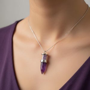 Könnte beinhalten: Eine silberne Halskette mit einem Anhänger aus einem lila Amethystkristall. Der Anhänger hat einen zylindrischen oberen Teil, der mit kleineren lila Kristallen gefüllt ist, und eine spitz zulaufende Amethystbasis. Die Halskette wird von einer Person getragen, die ein lila Oberteil trägt.