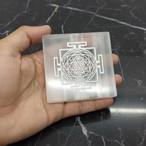 Könnte beinhalten: Ein quadratischer, durchscheinender Selenitkristall mit einem detaillierten, geätzten geometrischen Design. Das Design ist ein symmetrisches Muster aus Linien und Formen, möglicherweise ein heiliges Symbol. Der Kristall wird in einer Hand gehalten.