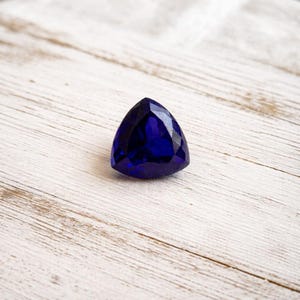 Könnte beinhalten: Ein facettierter, dreieckiger, tiefblauer Edelstein. Der Edelstein liegt auf einer hellen, strukturierten Holzoberfläche. Die Facetten des Edelsteins fangen das Licht ein und erzeugen Glanzlichter und Schatten.
