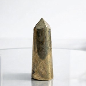Könnte beinhalten: Ein goldfarbener Kristallpunkt mit facettierter Spitze. Der Kristall hat eine zylindrische Form mit leicht verjüngter Basis. Die Oberfläche weist natürliche Farb- und Texturvariationen auf. Höhe ca. 10 cm.
