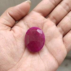 Könnte beinhalten: Ein facettierter, oval geformter, tief magentafarbener Edelstein. Der Stein wird in einer offenen Handfläche gehalten, wodurch seine polierte Oberfläche und die geometrischen Schnitte zur Geltung kommen. Die Farbe des Steins ist ein satter, gesättigter Farbton.