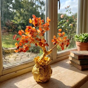 Könnte beinhalten: Dekorative Baumskulptur mit goldfarbenem Stamm und Sockel sowie orange-roten Edelsteinblättern. Der Baum steht in einem goldfarbenen Topf mit strukturierter Oberfläche auf einer Holzfensterbank. Eine Sukkulente im Topf und gestapelte Bücher sind ebenfalls zu sehen.