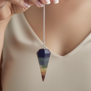 Könnte beinhalten: Ein facettierter Pendel mit einer silbernen Kette. Der Pendel hat einen Farbverlauf, beginnend mit Dunkelblau an der Spitze, über verschiedene Blau-, Grün-, Gelb- und Orangetöne bis hin zu einer braunen Spitze. Der Pendel wird von einer Person gehalten.
