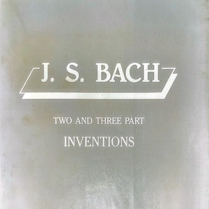Könnte beinhalten: Ein Buchcover mit dem Text "J. S. BACH" in Weiß, darunter die Worte "TWO AND THREE PART INVENTIONS". Der Hintergrund ist eine gedämpfte graue Farbe.