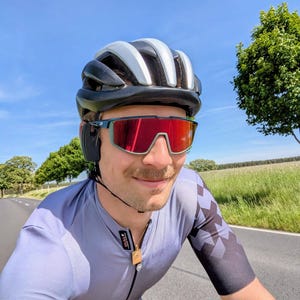 Könnte beinhalten: Ein Radfahrer trägt einen schwarz-weißen Helm, teal-farbene und rote Sonnenbrille und ein graues Radtrikot mit einem schwarz-grauen Rautenmuster auf den Ärmeln. Der Radfahrer fährt an einem sonnigen Tag auf einer asphaltierten Straße.