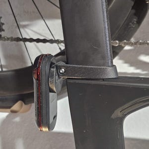 Könnte beinhalten: Ein schwarzes Garmin-Fahrradrücklicht ist an einem schwarzen Fahrradrahmen befestigt. Das Licht hat eine rote Linse und einen schwarzen Körper. Das Hinterrad und die Kette des Fahrrads sind im Hintergrund sichtbar. Das Licht ist an der Sattelstütze befestigt.