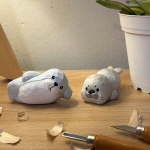 May include: Two hand-carved wooden seal figurines with a light blue and white painted finish. Each seal features a smooth, rounded body with visible carved facets, painted black eyes, a black nose, and small whiskers on a light-coloured surface.
