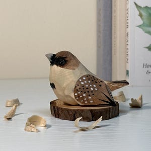 Puede incluir: Una figura de p&aacute;jaro tallada a mano en madera con cuerpo marr&oacute;n claro, marcas marr&oacute;n oscuro, una mancha negra en la cara y puntos blancos en las alas. El p&aacute;jaro est&aacute; posado sobre una peque&ntilde;a rodaja de madera natural rodeada de virutas de madera.