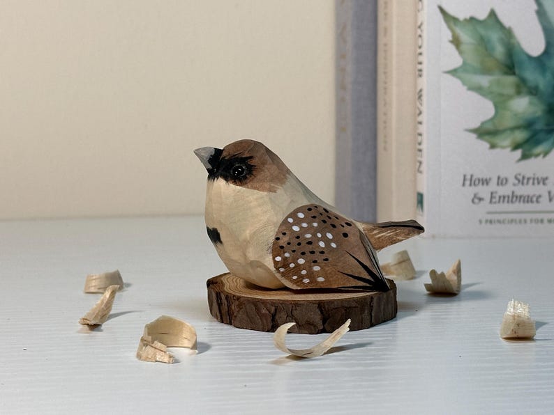 Puede incluir: Una figura de p&aacute;jaro tallada a mano en madera con cuerpo marr&oacute;n claro, marcas marr&oacute;n oscuro, una mancha negra en la cara y puntos blancos en las alas. El p&aacute;jaro est&aacute; posado sobre una peque&ntilde;a rodaja de madera natural rodeada de virutas de madera.