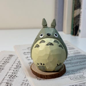 May include: A hand-carved wooden Totoro figurine, painted in shades of gray and cream, with black accents. The figure is perched on a small wooden slice, set against a backdrop of sheet music and books. The figurine is approximately 4 inches tall.