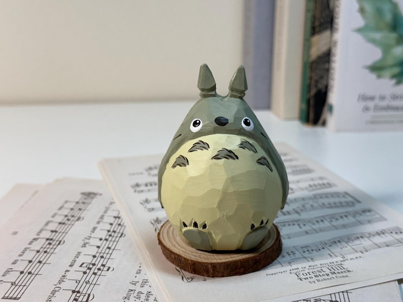 May include: A hand-carved wooden Totoro figurine, painted in shades of gray and cream, with black accents. The figure is perched on a small wooden slice, set against a backdrop of sheet music and books. The figurine is approximately 4 inches tall.