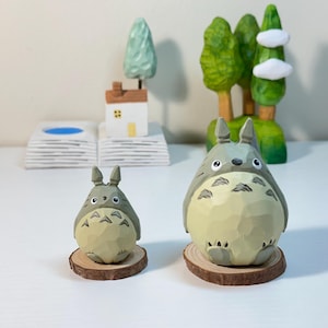 May include: Two wooden Totoro figurines, one large and one small, sit on wooden slices. The larger figure is in front of a carved wooden forest scene with trees and clouds. A small house and tree are in the background.