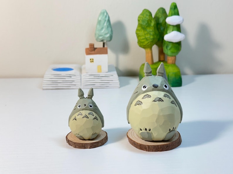 May include: Two wooden Totoro figurines, one large and one small, sit on wooden slices. The larger figure is in front of a carved wooden forest scene with trees and clouds. A small house and tree are in the background.