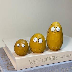 May include: Three whimsical, hand-carved wooden figures in varying sizes, each with a unique expression. The figures are painted in shades of yellow and brown, with white eyes and black pupils. They are displayed on a stack of books.