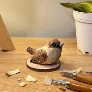 Puede incluir: Una figura de p&aacute;jaro de madera tallada a mano con tonos marrones y crema, detalles pintados en las alas y un ojo oscuro. El p&aacute;jaro est&aacute; montado sobre una peque&ntilde;a base circular de madera, cerca de herramientas de tallado y virutas de madera.