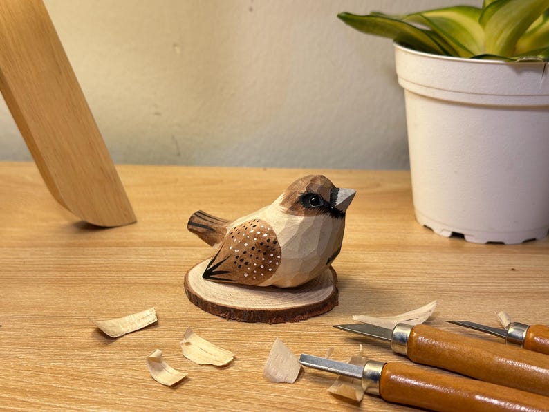 Puede incluir: Una figura de p&aacute;jaro de madera tallada a mano con tonos marrones y crema, detalles pintados en las alas y un ojo oscuro. El p&aacute;jaro est&aacute; montado sobre una peque&ntilde;a base circular de madera, cerca de herramientas de tallado y virutas de madera.