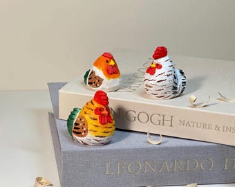 May include: Three hand-painted wooden rooster figurines in vibrant colors. One is yellow and orange, another is white with black stripes, and the third is orange and white. They are perched on books with gold lettering.