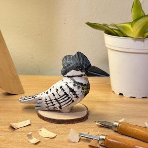 Puede incluir: Una figura de madera tallada a mano de un mart&iacute;n pescador con cresta gris oscuro, pico negro y plumaje blanco con marcas negras. El ave est&aacute; montada sobre una peque&ntilde;a base circular de madera con textura de veta visible.