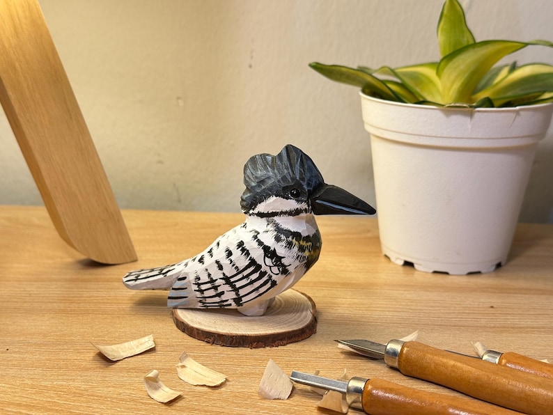 Puede incluir: Una figura de madera tallada a mano de un mart&iacute;n pescador con cresta gris oscuro, pico negro y plumaje blanco con marcas negras. El ave est&aacute; montada sobre una peque&ntilde;a base circular de madera con textura de veta visible.