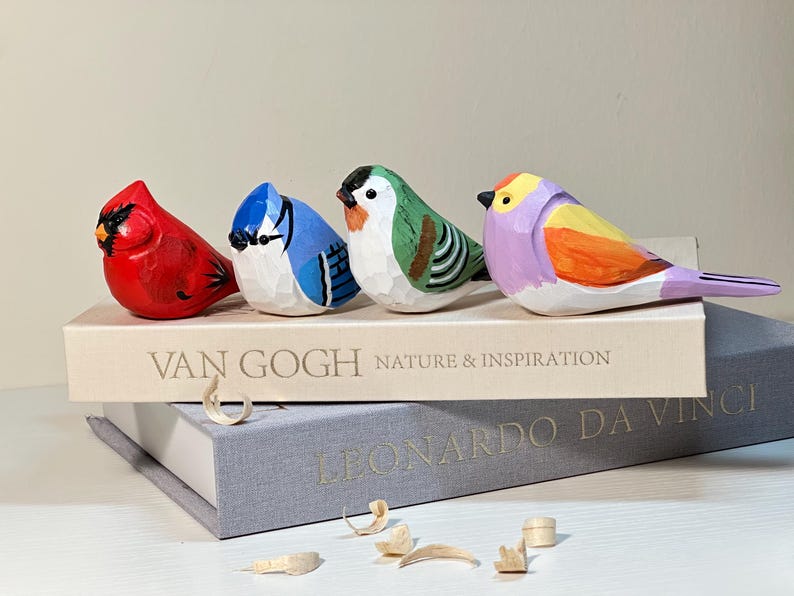 Puede incluir: Cuatro figuras de p&aacute;jaros de madera talladas a mano en rojo, azul, verde y morado, sobre un libro titulado VAN GOGH NATURE & INSPIRATION, que descansa sobre un libro gris titulado LEONARDO DA VINCI, con virutas de madera en el primer plano.
