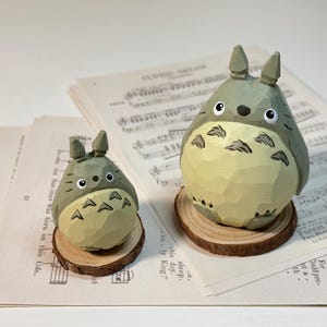 May include: Two wooden Totoro figurines, one large and one small, sit on wooden slices. The figures are gray with cream-colored bellies and black eyes. They rest on sheet music. The larger Totoro is approximately 4 inches tall.