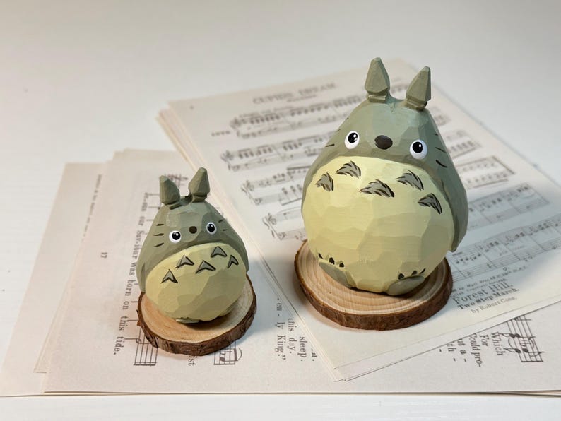 May include: Two wooden Totoro figurines, one large and one small, sit on wooden slices. The figures are gray with cream-colored bellies and black eyes. They rest on sheet music. The larger Totoro is approximately 4 inches tall.