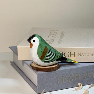 Puede incluir: Una figura de p&aacute;jaro de madera tallada a mano con espalda verde, pecho blanco, marcas negras en la cabeza y detalles naranjas cerca del pico. El p&aacute;jaro est&aacute; posado sobre una peque&ntilde;a base de rodaja de madera, mostrando texturas talladas visibles.