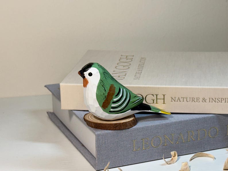Puede incluir: Una figura de p&aacute;jaro de madera tallada a mano con espalda verde, pecho blanco, marcas negras en la cabeza y detalles naranjas cerca del pico. El p&aacute;jaro est&aacute; posado sobre una peque&ntilde;a base de rodaja de madera, mostrando texturas talladas visibles.