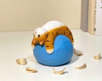 Puede incluir: Figura de madera tallada a mano de un gato descansando sobre una bola azul. El gato es blanco y naranja con rayas negras. La bola es de color azul brillante. Virutas de madera esparcidas alrededor.