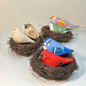 Puede incluir: Una colecci&oacute;n de seis figuras de aves de madera talladas a mano, cada una descansando en un peque&ntilde;o nido circular de ramitas. Las aves presentan varios detalles pintados, incluyendo un cardenal rojo brillante, un arrendajo azul, un ave verde y tonos madera.