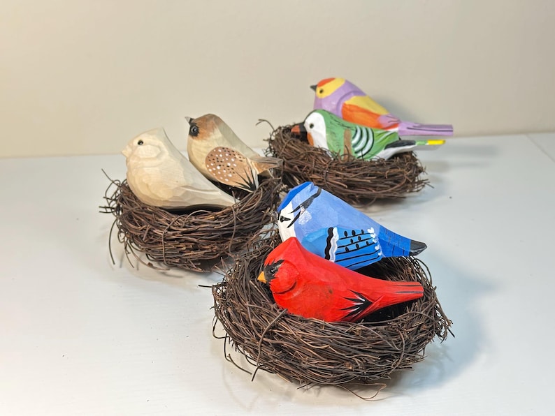 Puede incluir: Una colecci&oacute;n de seis figuras de aves de madera talladas a mano, cada una descansando en un peque&ntilde;o nido circular de ramitas. Las aves presentan varios detalles pintados, incluyendo un cardenal rojo brillante, un arrendajo azul, un ave verde y tonos madera.
