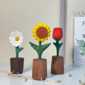 Może przedstawiać: Trzy drewniane rzeźby kwiatów: biała stokrotka, żółty słonecznik i czerwona róża. Każdy kwiat umieszczony jest w brązowej drewnianej doniczce. Słonecznik ma brązowy środek. Zielone liście i łodygi podtrzymują kwiaty. Wióry drzewne są rozrzucone.