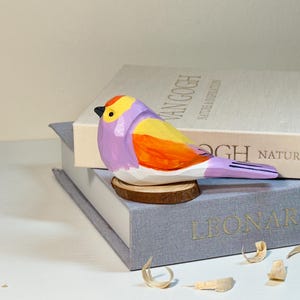 Puede incluir: Una figura de p&aacute;jaro tallada a mano en madera con una vibrante paleta de colores morado, amarillo y naranja, con un peque&ntilde;o pico negro. El p&aacute;jaro est&aacute; posado sobre una base circular de madera, mostrando facetas talladas y un acabado pintado.