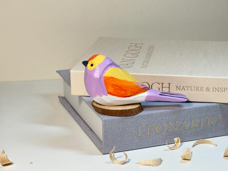 Puede incluir: Una figura de p&aacute;jaro tallada a mano en madera con una vibrante paleta de colores morado, amarillo y naranja, con un peque&ntilde;o pico negro. El p&aacute;jaro est&aacute; posado sobre una base circular de madera, mostrando facetas talladas y un acabado pintado.
