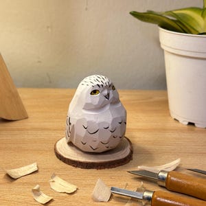 Puede incluir: Una figura de b&uacute;ho nival tallada a mano en madera con acabado blanco, que presenta superficies facetadas, detalles de plumas pintados en negro y ojos amarillos. El b&uacute;ho est&aacute; montado sobre una base de rodaja de madera circular.