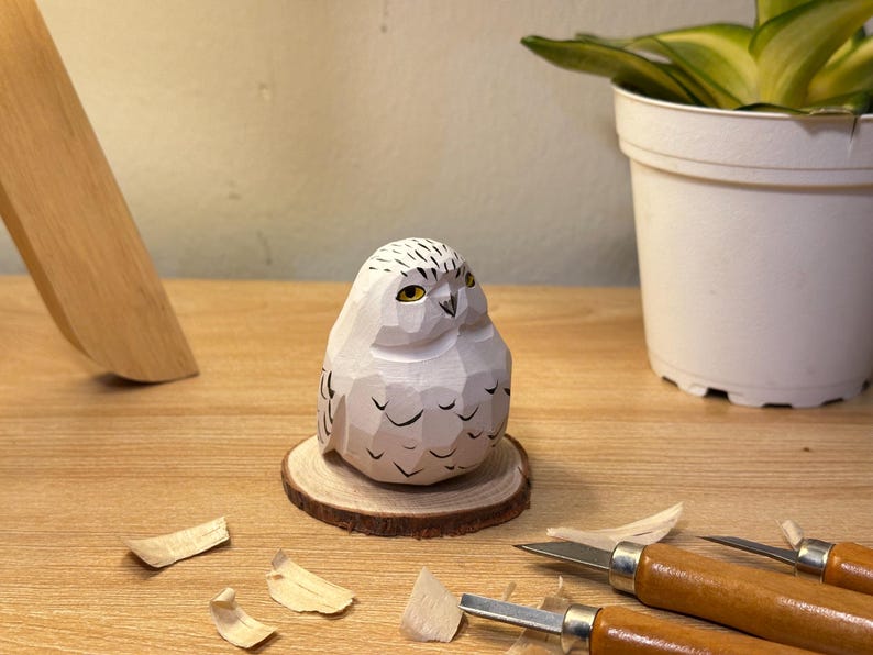 Puede incluir: Una figura de b&uacute;ho nival tallada a mano en madera con acabado blanco, que presenta superficies facetadas, detalles de plumas pintados en negro y ojos amarillos. El b&uacute;ho est&aacute; montado sobre una base de rodaja de madera circular.