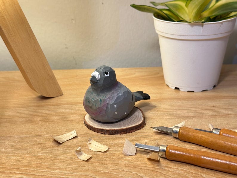 Puede incluir: Una figura de paloma tallada a mano en madera con cuerpo gris, marcas blancas en el pico y detalles negros en los ojos. El ave est&aacute; montada sobre una peque&ntilde;a base circular de rodaja de madera, con facetas visibles del proceso de tallado.