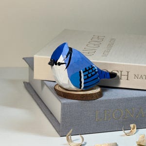 Puede incluir: Una figura de arrendajo azul tallada a mano en madera con un acabado pintado en azul vibrante, blanco y negro. El p&aacute;jaro est&aacute; posado sobre una peque&ntilde;a base circular de rodaja de madera natural, mostrando facetas talladas y un pico negro.