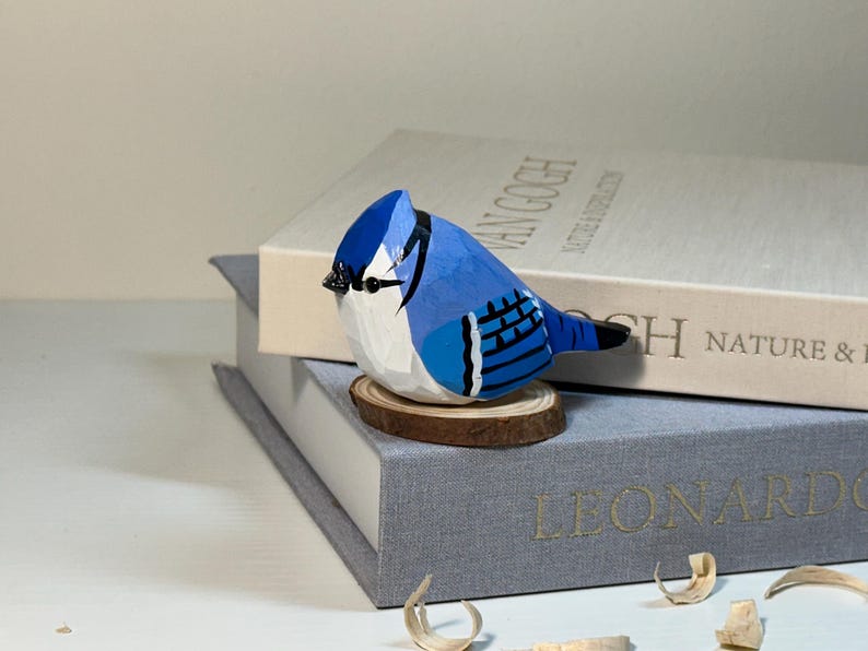 Puede incluir: Una figura de arrendajo azul tallada a mano en madera con un acabado pintado en azul vibrante, blanco y negro. El p&aacute;jaro est&aacute; posado sobre una peque&ntilde;a base circular de rodaja de madera natural, mostrando facetas talladas y un pico negro.