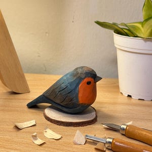 Puede incluir: Una figura de petirrojo tallada a mano en madera, con el dorso y las alas de color azul gris&aacute;ceo oscuro y el pecho de color naranja brillante. El p&aacute;jaro est&aacute; montado sobre una peque&ntilde;a base circular de madera clara, con texturas talladas y un ojo negro.
