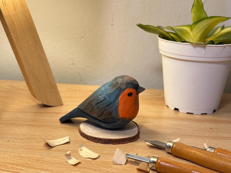 Puede incluir: Una figura de petirrojo tallada a mano en madera, con el dorso y las alas de color azul gris&aacute;ceo oscuro y el pecho de color naranja brillante. El p&aacute;jaro est&aacute; montado sobre una peque&ntilde;a base circular de madera clara, con texturas talladas y un ojo negro.