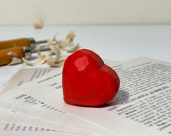 Puede incluir: Un vibrante corazón rojo de madera tallada descansa sobre páginas de recetas antiguas. El corazón, con su diseño facetado, resalta su color. Herramientas de tallado y virutas de madera en el fondo sugieren un objeto artesanal.