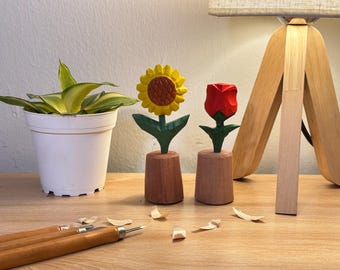 Puede incluir: Dos figuras de flores de madera talladas a mano, un girasol amarillo y un tulipán rojo, cada una montada sobre una base cilíndrica de madera. Las flores presentan tallos y hojas verdes, con texturas talladas visibles en los pétalos.