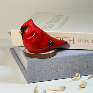 Puede incluir: Una figura de cardenal tallada a mano en madera, pintada en rojo vibrante con marcas faciales negras y un pico amarillo. El p&aacute;jaro est&aacute; posado sobre una peque&ntilde;a base circular de rodaja de madera natural, mostrando texturas de vetas y plumas.