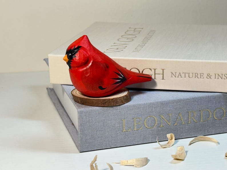 Puede incluir: Una figura de cardenal tallada a mano en madera, pintada en rojo vibrante con marcas faciales negras y un pico amarillo. El p&aacute;jaro est&aacute; posado sobre una peque&ntilde;a base circular de rodaja de madera natural, mostrando texturas de vetas y plumas.