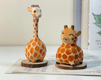 Puede incluir: Dos figuras de jirafa de madera talladas a mano. Una es alta con cuello largo, la otra es corta y robusta. Ambas tienen patrones de manchas naranjas y blancas y se asientan sobre pequeñas bases de madera. Las figuras de jirafa son decorativas.