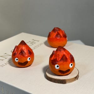 May include: Three orange and red wooden figures, resembling flames, are arranged on a book. Each figure has a smiling face with white eyes and a dark brown mouth. One figure rests on a small wooden disc.