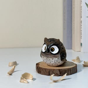 Puede incluir: Una peque&ntilde;a figura de b&uacute;ho tallada a mano en madera con detalles pintados en marr&oacute;n y blanco, que presenta grandes ojos con borde blanco, iris amarillos y pupilas negras, posada sobre una rodaja de madera circular con corteza natural.
